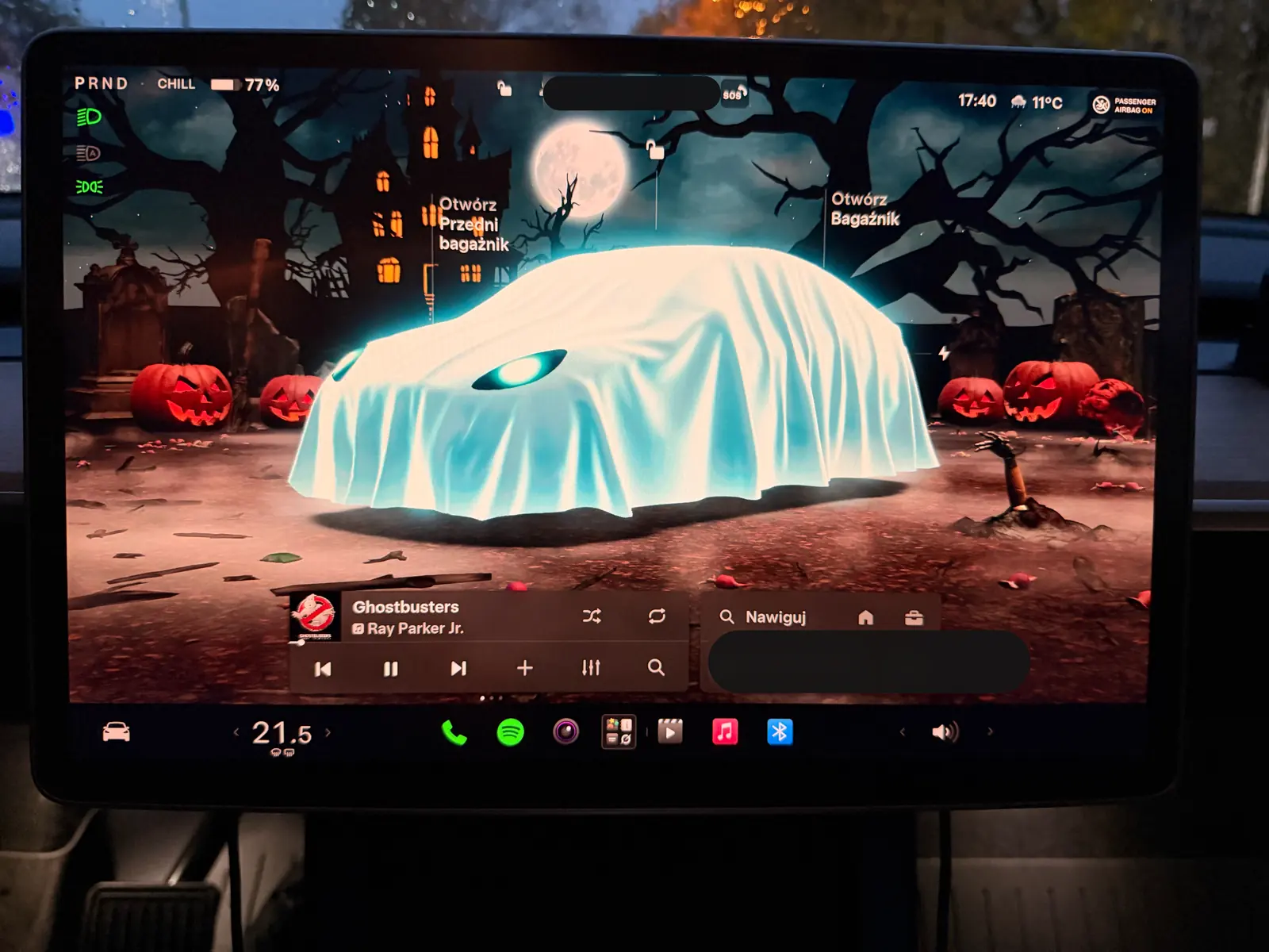 Ekran Tesli w trybie Halloween: auto pod białym prześcieradłem-duchem na tle nocy, pełni, nawiedzonego domu i dyń
